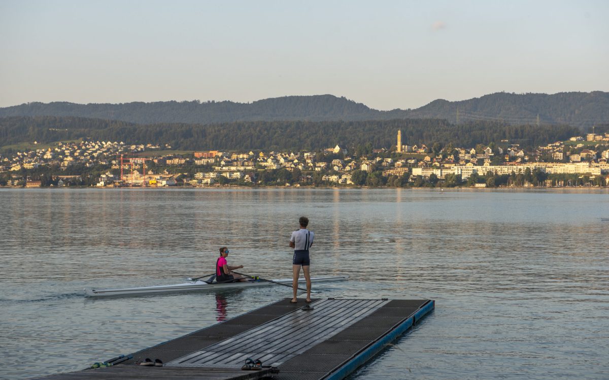 Coach steht auf Steg und spricht mit Ruderin im Einerboot auf dem See bei Sonnenaufgang